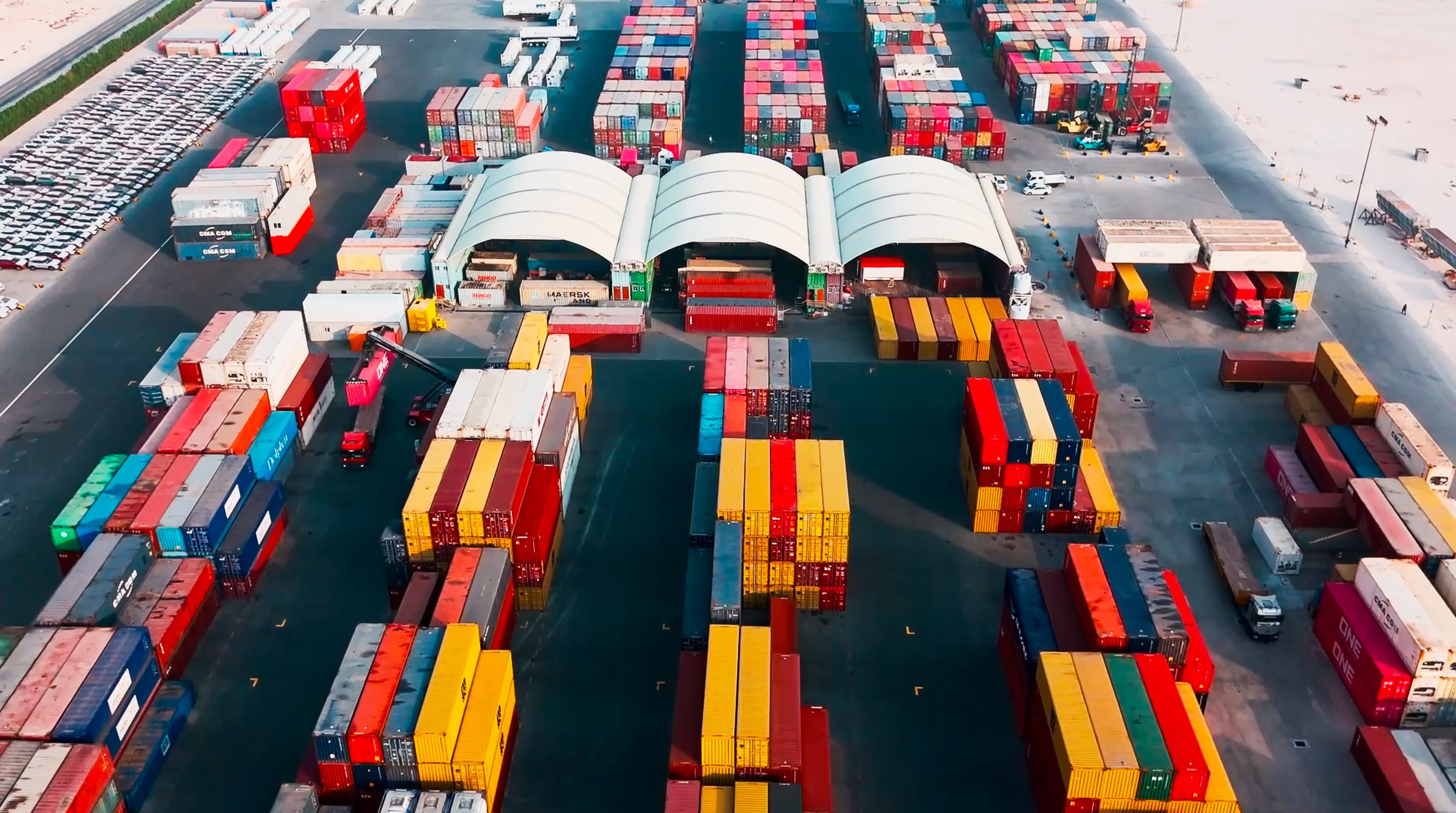 Aerial view of a busy shipping port with rows of colorful shipping containers and three large white tents. Trucks maneuver around the organized stacks.